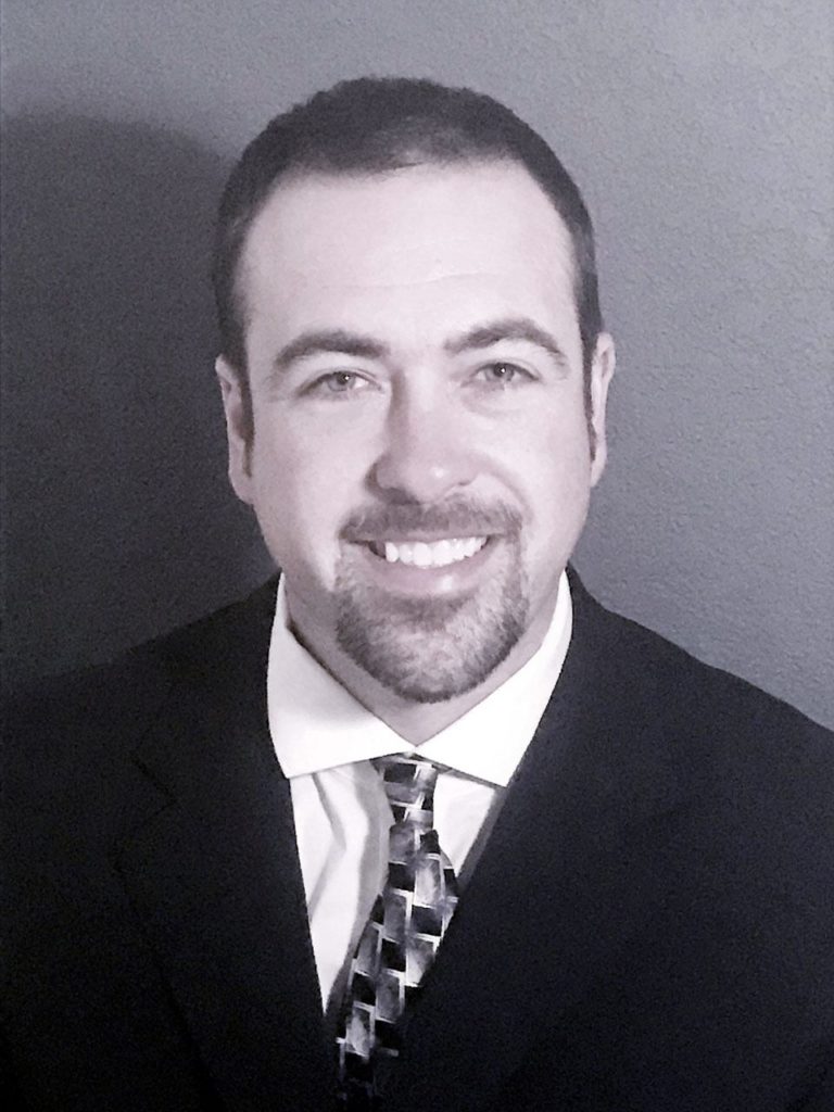 headshot photograph of Shawn Casey wearing a suit and tie, smiling in front of a gray background